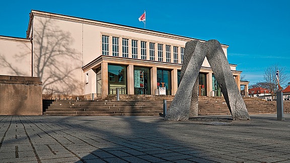 Theater Zittau, ein quadratischer Bau mit Eingangsbereich und darauf hinführenden Stufen, davor eine abstrakte Plastik.