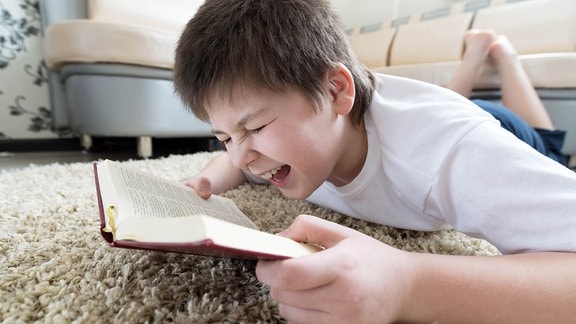 Ein lachender Junge liegt mit einem Buch auf dem Wohnzimmerboden
