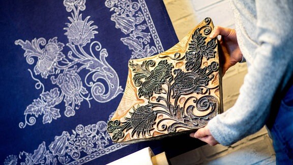 Blaudruckerin Sabrina Schuhmacher hält in ihrer Werkstatt einen historischen Druckstock, eine sogenannte Model, vor einen blau gefärbten Stoff mit dem gleichen Muster.