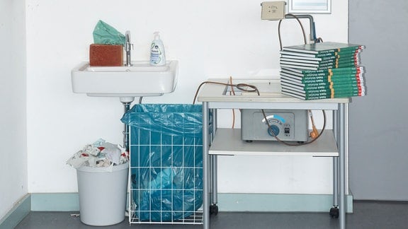 Aus der Serie "Soziale Gerechtigkeit" von Fotografin Susanne Keichel: Ein Klassenzimmer, unter einem Waschbecken stehen zwei Mülleimer, daneben ein Overheadprojektor mit einem Stapel Bücher. 