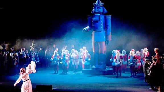 Eine Szene aus einem Theaterstück: Um einen riesigen Nussknacker steht ein Chor aus Weihnachtsmännern. Eine Frau im Kleid schaut erstaunt zu.
