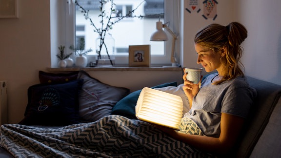 Junge Frau auf Sofa hält beleuchtetes Buch und Teetasse