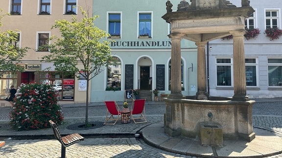 Der Platz vor der Robert-Philipp-Buchhandlung in Kamenz. Zwei rote Klappstühle stehen in der Mitte des Bildes neben einem altertümlichen Brunnen. Daneben sind zwei Bäume und ein Blüten versehener Busch zu sehen. Im Hintergrund ist eine Häuserfassade, in deren Mitte die Buchhandlung zu sehen ist. 