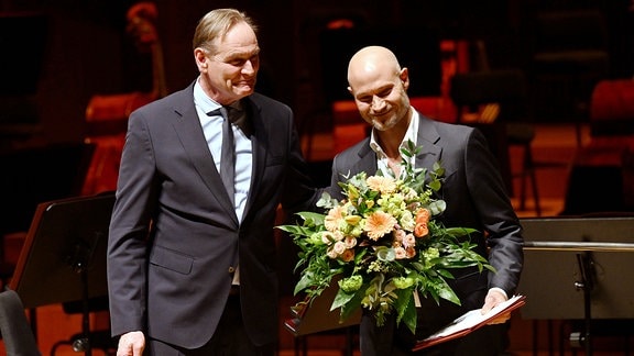 Zwei Männer im Anzug stehen auf einer Bühne; einer hält einen Blumenstrauß und eine Urkunde in der Hand.