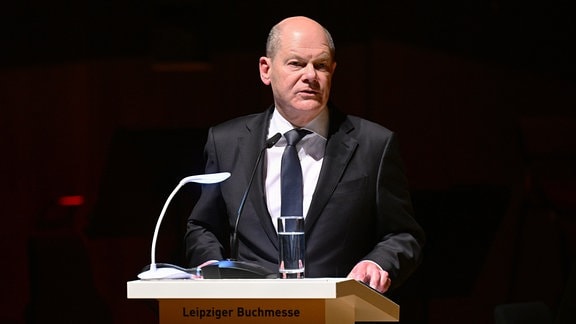 Bundeskanzler Olaf Scholz: ein Mann in dunklem Anzug, weißem Hemd und Krawatte steht vor einem dunklen Hintergrund und spricht in ein Mikrofon.