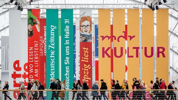 eine Gruppe von Menschen läuft über eine Brücke in einem verglasten Gebäude; im Hintergrund bunte Werbebanner