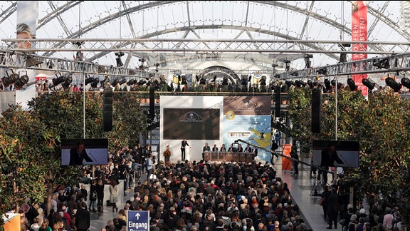 Blick in die Glashalle auf der Leipziger Messe: in der Mitte sitzen zahlreiche Menschen und blicken auf eine Bühne
