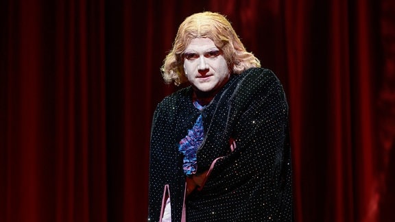 Ein Mann mit lankgen blonden Haaren und weiß geschminkten Gesicht steht vor einem roten Vorhang.