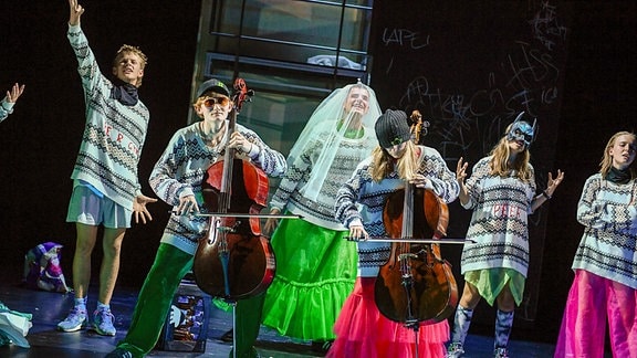 Zahlreiche Menchen in übergroßen Pullovern, bunten Hosen und Röcken stehen auf einer Bühne, zwei spielen stehend Cello.