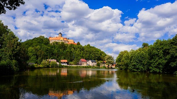 Burg Schönfels thront auf einem umwaldeten Fels über einem Fluss. Im Hintergrund: strahlend blauer Himmel mit kleinen Wolken.