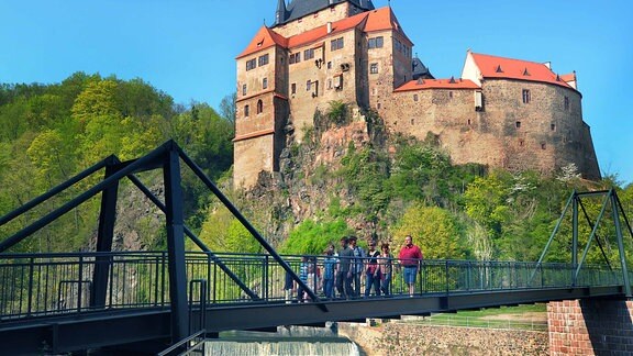 Eine Metallbrücke über einen Fluss, im Hintergrund ist Burg Kriebstein zu sehen. 