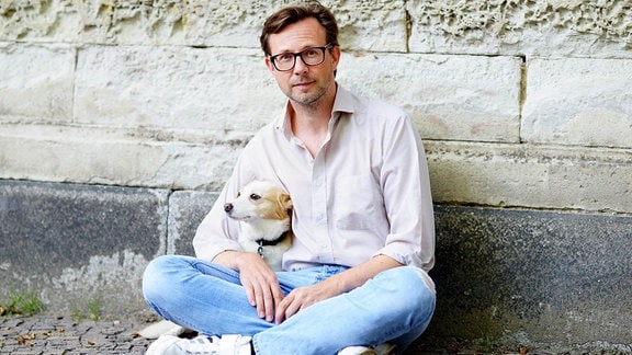 Carl-Christian Elze, ein Mann mit Jeans, Hemd und Brille sitzt an einer Mauer und umarmt einen Hund.