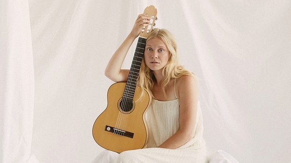 Eine Frau sitzt mit einer Holzgitarre vor einer mit Laken verhängten Wand