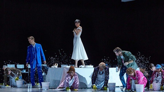 Eine Frau in weißem Kleid steht auf einem Tisch. Neben ihr ein Mann in blauen Anzug. Auf dem Boden hocken weitere Personen.