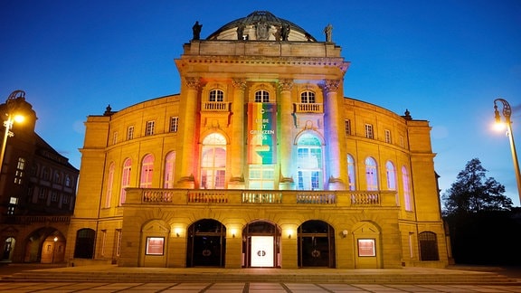 Theater Chemnitz: Ein historisches Gebäude in Abendstimmung ist mit Regenbogenfarben beleuchtet.
