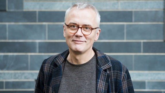 Ein Mann mit kurzem weißen Haar und Brille steht vor einer grau gefliesten Wand  