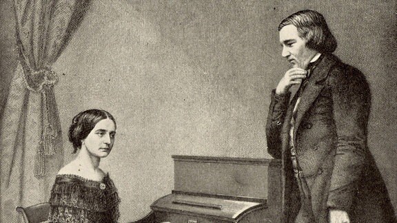 Robert Schuhmann steht neben einem Klavier, an dem seine Frau Clara Schuhmann spielt.
