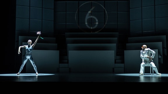 Zwei Personen in futuristischen Kostümen stehen auf einer sonst dunklen Bühne in Posen in Lichtkegeln.
