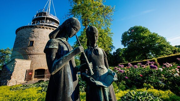 Skultptur zweier Mädchen vor Blumenrabatten und einem Turm