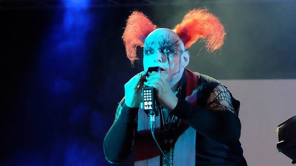 Ein kostümierter Bruno Kramm mit orangefarbenen Clowns-Haaren auf einer Bühne.