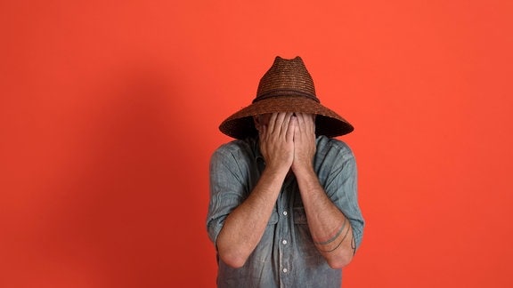 Ein Mann hält sich die Hände vor das Gesicht und trägt einen Hut.
