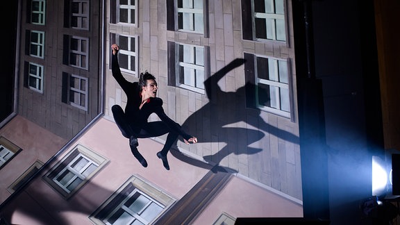Szene aus dem Theaterstück "Der Meister und Margarita" in Weimar: Ein Mann springt mit angewinkelten Beinen in die Höhe, im Hintergrund eine Häuserfassade.