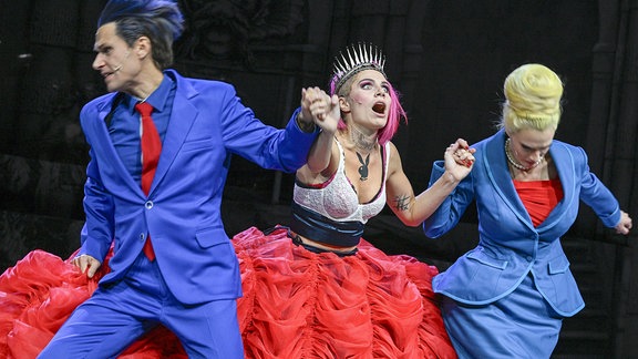 Eine Frau mit einem ausladenden roten Rock wird von zwei Personen in hellblauen Anzügen an den Händen gehalten. 