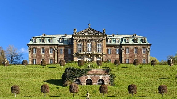 Auf einem begrünten Hügel mit angelegten Terassen erhebt sich Schloss Burgscheidungen, ein Gebäude mit verzierter Fassade und hellem Dach. 