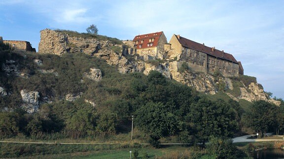 Auf einem Hügel erhebt sich Burg Wendelstein hinter Felsen, im Vordergrund ein Fluss und begrüntes Ufer.  
