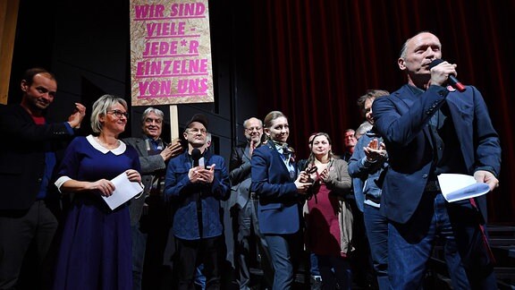 Hasko Weber spricht 2019 auf der Bühne, im Hintergrund stehen mehrere Menschen und halten ein Schild hoch, auf dem steht: "Wir sind viele. Jede:r einzelne von uns."