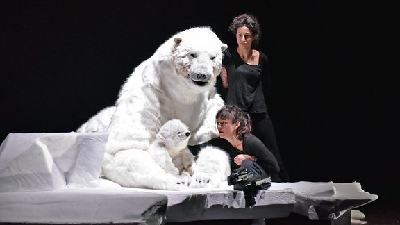 Eine Person in Schwarz sitzt auf einem weißen Sofa mit Puppen eines großen und eines kleinen Eisbären. Dahinter steht eine weitere Person in Schwarz.