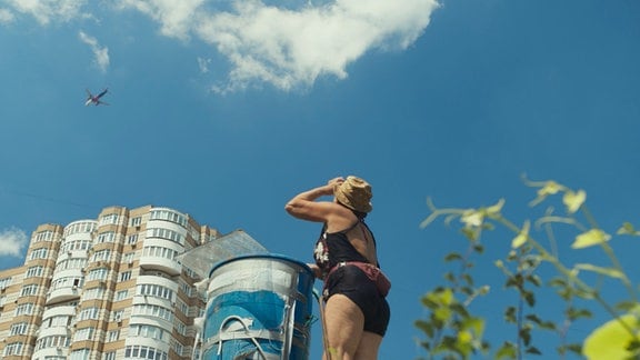 Filmstill von "Flowers of Ukraine", eine Frau schaut auf ein Hochhaus und blauen Himmel