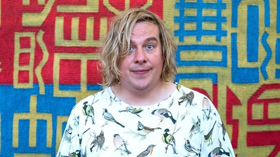 Ein Mann mit blonden Haaren und einem T-Shirt mit aufgedruckten Vögeln steht vor einer bunt gemusterten Wand