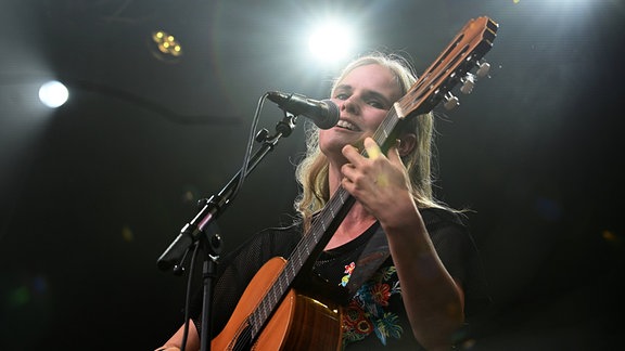 Eine Frau mit blonden langen Haaren mit Gitarre in der Hand singt lächelnd auf einer erleuchteten Bühne