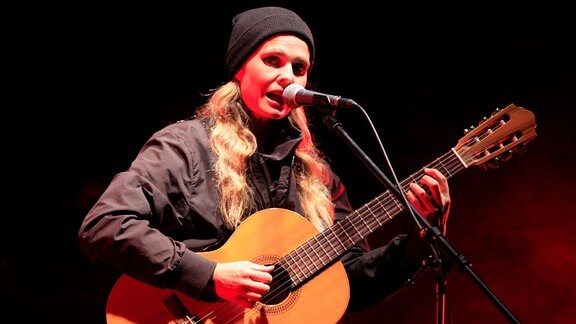 Dota Kehr: eine Frauen mit blonden Haaren und Mütze steht auf der Bühne und singt mit der Gitarre in der Hand.