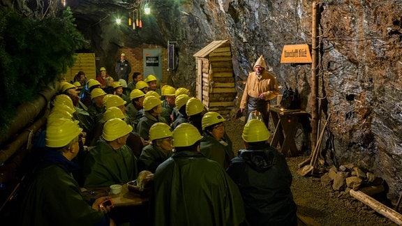 Besucherinnen und Besucher in einem Bergwerk: Die Anwesenden tragen Schutzhelme und lauschen dem Vortrag eines Bergmannes
