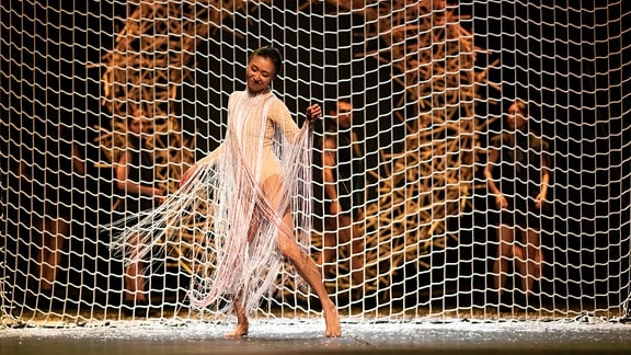 Eine Frau in einem weißen Netzkleid tanzt vor einem vor einem grobmaschigen Netz