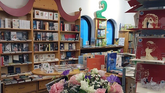 Blick in den Bücherladen Fabularium Magdeburg. Zu sehen sind verschiedene Bücherregale in einem Zimmer. In der Mitte hängt ein Kronleuchter, im Vordergrund steht ein Tisch mit Büchern und einem großen Blumenstrauß.