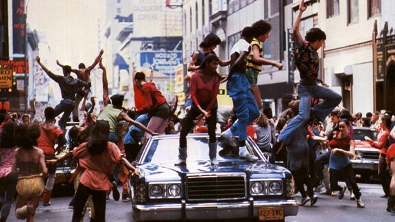 Junge Menschen tanzen auf einem Auto auf der Straße - Filmausschnit aus "Fame - Der Weg zum Ruhm" (1980).