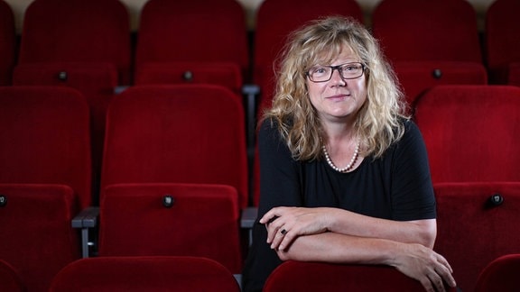 Eine Frau mit lockigen blonden Haaren und Brille sitzt in einer Reihe roter Theaterklappsessel