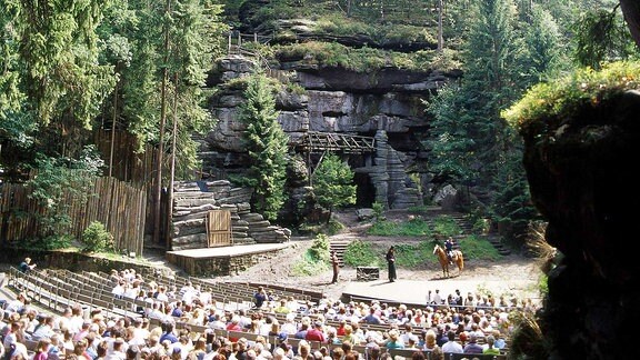 Besucher bei einer Aufführung in der Felsenbühne Rathen, man sieht die gefüllten Besucherreihen, vorne die Felsenbühne und Felsen, die in den blauen Himmel ragen