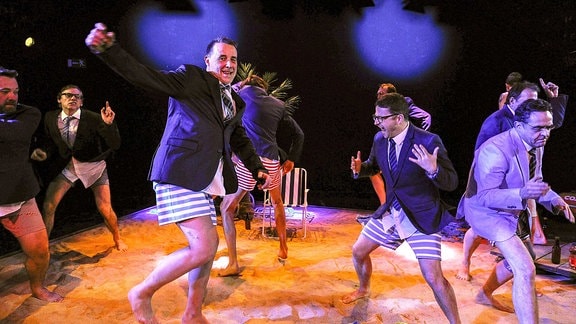 Menschen in gestreiften Badehosen und Anzugsjacken tanzen wild über einen künstlichen Sandstrand auf einer Bühne.