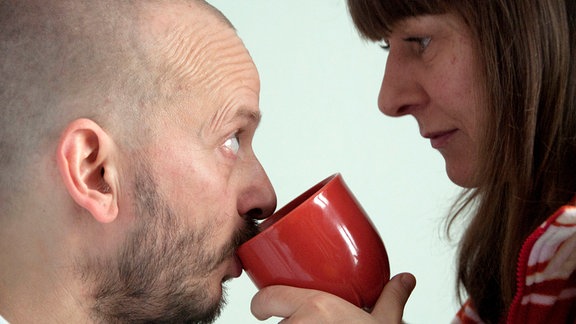 Eine Frau hilft ihrem Mann beim Trinken aus einer Tasse, beide blicken sich in die Augen
