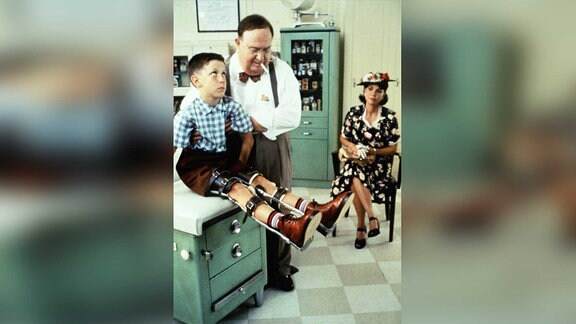 Der junge Forrest Gump mit Beinschinen. Der Arzt hilft ihm beim Aufstehen. Seine Mutter im Hintergrund.