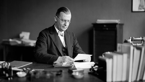 Historisches Foto: Ein Mann in Anzug sitzt an einem Schreibtisch und liest einen Brief.