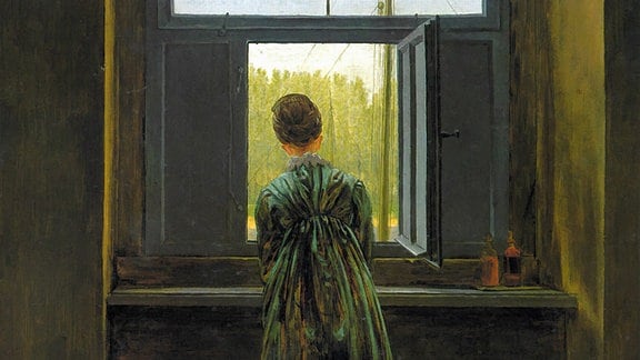 Das Gemälde "Frau am Fenster" von Caspar David Friedrich zeigt eine Frau, die mittig vor einem Fenster steht, sie trägt ein grünes Kleid.