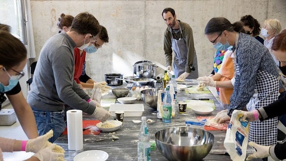 Ein Mann mit Brille, schwarzem lichten Haar und schwarzem Bard steht mit anderen Menschen an einem Tisch. Er zeigt ihnen wie man das jüdische Gericht Kreplach kocht. Das sind gefüllte Kartoffeln-Teigtaschen.
