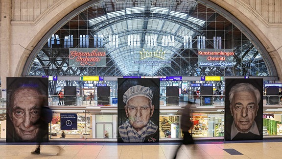 Reisende gehen an großformatigen Fotos von Überlebenden der NS-Verfolgung in der Ausstellung „Gegen das Vergessen“ auf dem Hauptbahnhof Leipzig vorbei