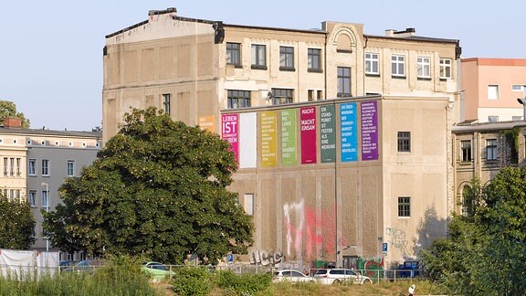 Blick durch eine Bachlandschauft auf einen Gebäude, an dem mehrere bunte Banner hängen.