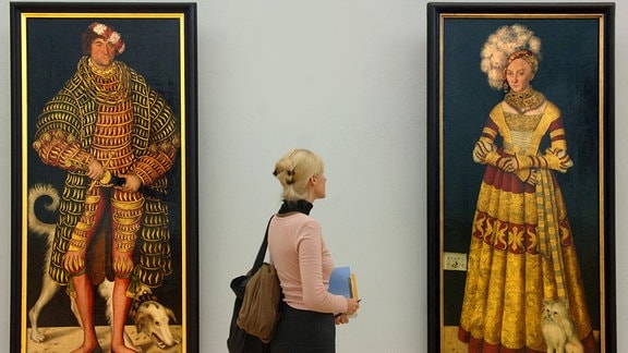 Eine junge Frau steht zwischen den Bildern "Herzog Heinrich der Fromme" und "Herzogin Katharina von Mecklenburg" und bertrachtet die Frau